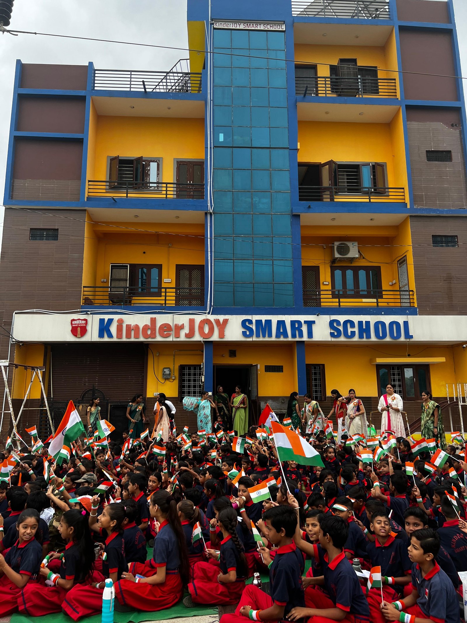 Kinder Joy Smart School students and staff waving the Indian tricolor during Independence Day celebrations outside the school building